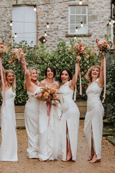 Bride in Alexandra Grecco dress with bridesmaids in white one shoulder dresses and neutral wedding bouquets.