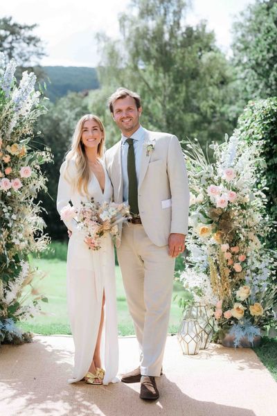 Bride wearing custom Bon Bride gown with plunging neckline and front slit with groom in cream suit in front of pastel flower displays for garden tipi wedding 