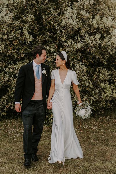 Bride in satin Ghost wedding dress walking with the groom in a morning suit on their wedding day.