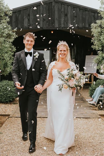 Bride in handmade wedding dress with crown walking with groom in black tie. 