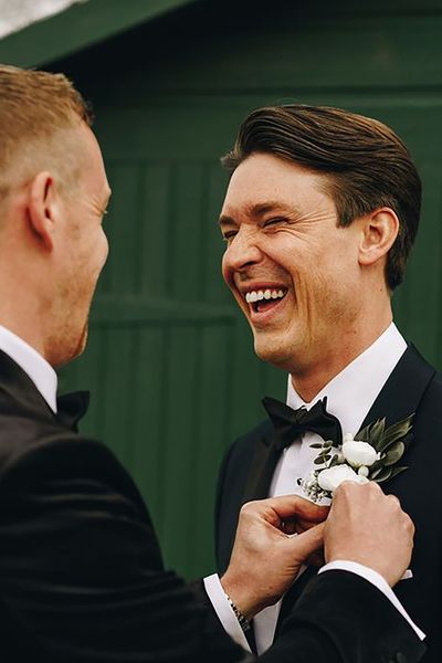 groom and his best man wearing black tie wedding suits while best man helps groom with buttonhole