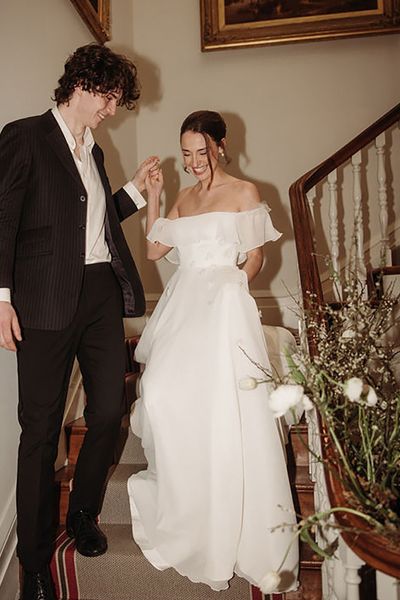 bride coming down the stairs with groom while wearing off-shoulder maxi wedding dress