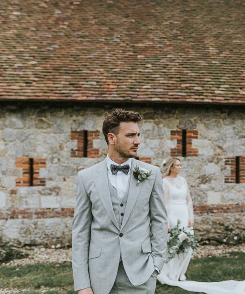 Portrait of the groom in a grey three-piece wedding suit with his bride in the background