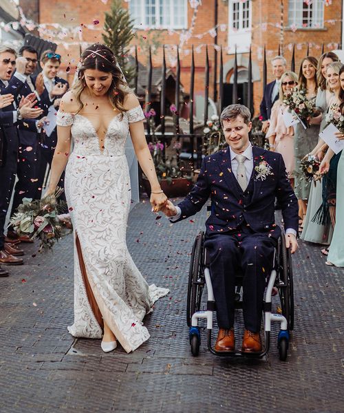 Bride in fitted wedding dress has confetti exit with groom in a blue suit in a wheelchair.