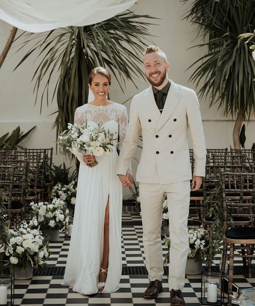 bride in lace Catherine Dean wedding dress with white wedding bouquet stands with husband in beige suit at Gunnesbury Park Orangery wedding altar