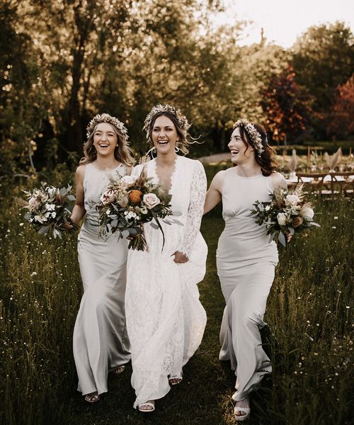 Bridesmaids in silver satin dresses and bride in a lace dress walking through the fields with a dried flower crown on their heads