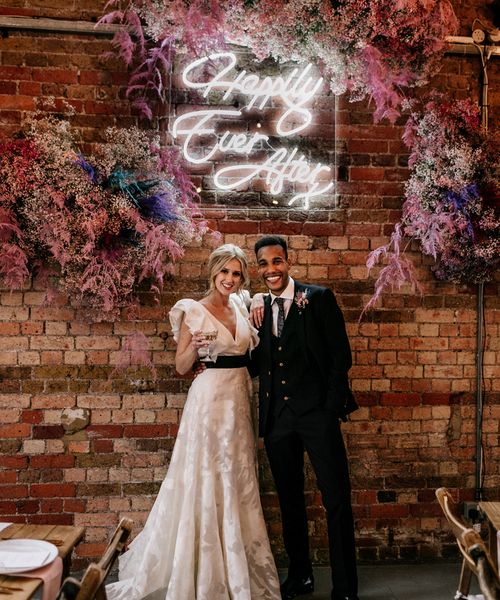 Prezola wedding couple standing in front of a neon happily ever after sign surround by pink gypsophila and wearing a ruffle sleeve wedding dress