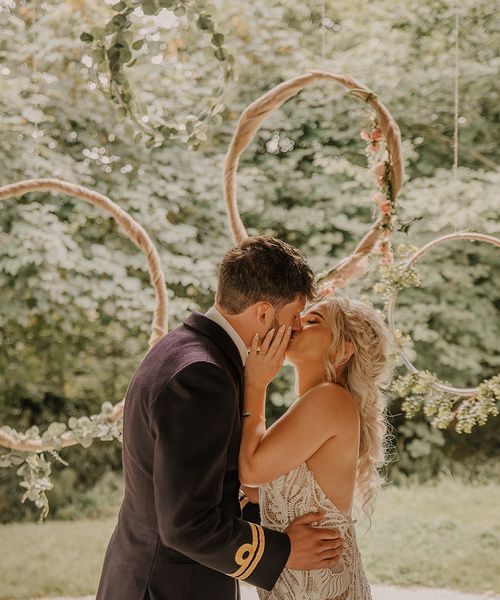 Hula hoop decor for the altar with the bride in a boho lace wedding dress kissing the groom in military uniform. 