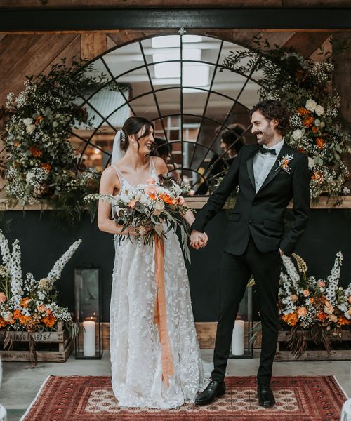 bride in a lace dress and groom in a black tuxedo at new Lake District wedding venue with oval micro back drop and colourful floral arrangements 