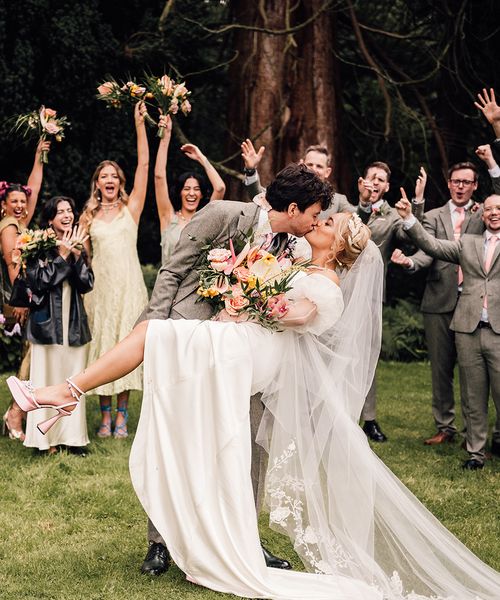 Groom dips bride in pink wedding shoes for a kiss with wedding party cheering in the background