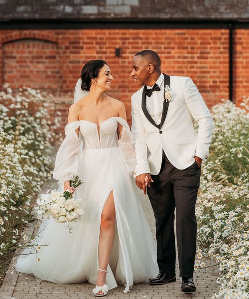 bride in off-the-shoulder tulle wedding dress and groom in black and white tuxedo walk hand in hand through white flowers at Crumplebury