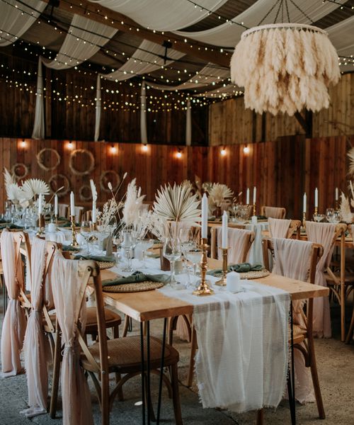 Romantic East Yorkshire Barns wedding reception with pink chiffon chair back decor, pampas grass chandelier, fairy light backdrop and dried palm leaf centrepieces 