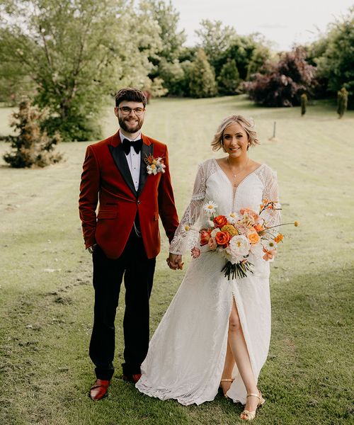 Bride holding an orange and yellow wedding flower bouquet in a boho dress with groom in an orange tux