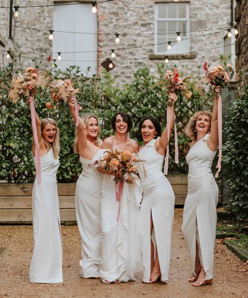 Bride in Alexandra Grecco dress with bridesmaids in white one shoulder dresses and neutral wedding bouquets.