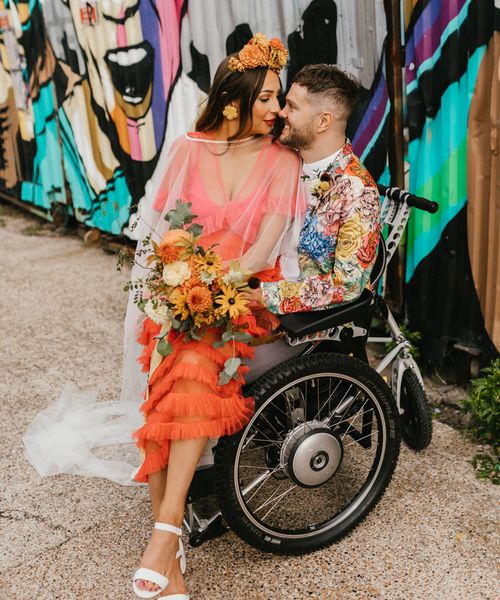 Bride in a coral wedding dress sitting on her grooms lap in a wheelchair for brightly coloured intimate elopement