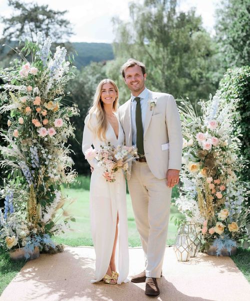 Bride wearing custom Bon Bride gown with plunging neckline and front slit with groom in cream suit in front of pastel flower displays for garden tipi wedding 