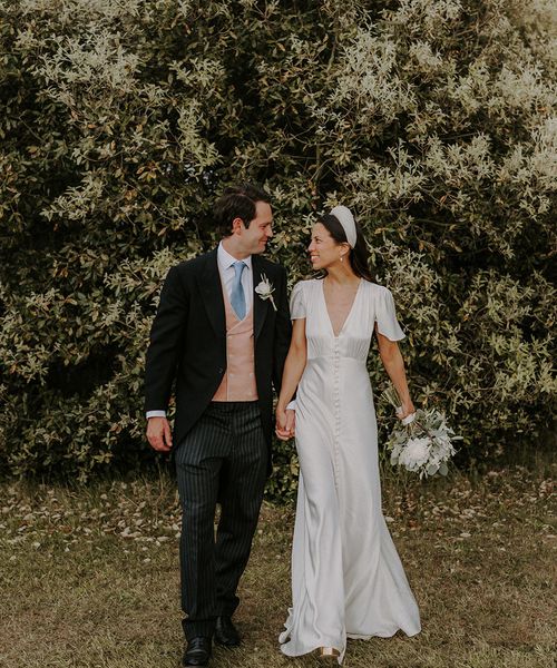 Bride in satin Ghost wedding dress walking with the groom in a morning suit on their wedding day.