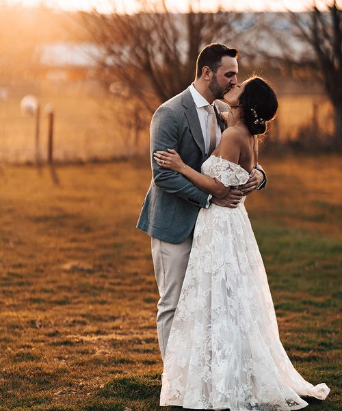 Bride in floral lace wedding dress with groom in grey suit share a kiss before their mariachi band wedding entertainment.