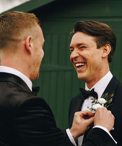 groom and his best man wearing black tie wedding suits while best man helps groom with buttonhole