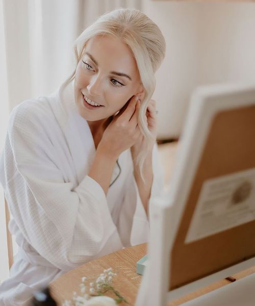 Bride in white waffle robe with blonde hair in an updo with radiant, glowy skin and makeup