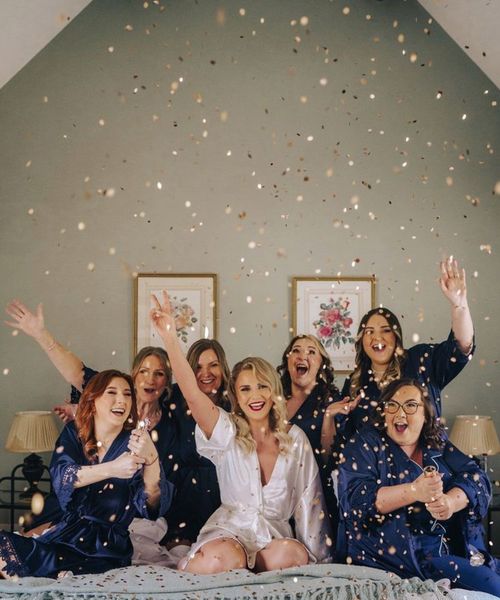 Bridesmaids in satin navy robes and pyjamas popping gold confetti canon as they get ready together