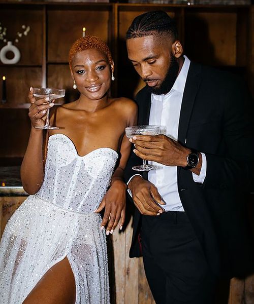 Bride in sparkly sweetheart strapless dress and leg slit holding a glass of champagne, standing with groom in skinny black suit for cocktail hour at wedding