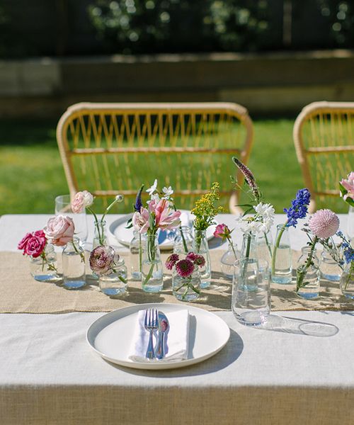 diy wedding centerpiece with mini glass vases and bottles and single stems of brightly coloured spring flowers on a linen tablecloth