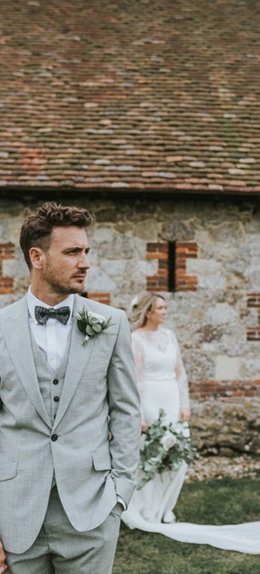 Portrait of the groom in a grey three-piece wedding suit with his bride in the background