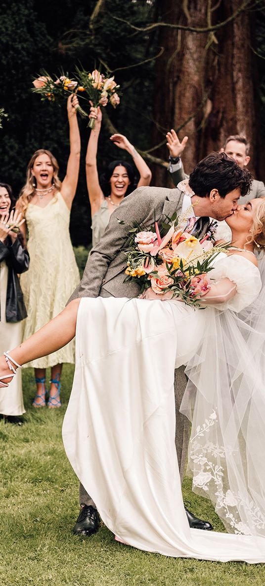 Groom dips bride in pink wedding shoes for a kiss with wedding party cheering in the background