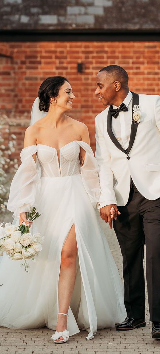 bride in off-the-shoulder tulle wedding dress and groom in black and white tuxedo walk hand in hand through white flowers at Crumplebury