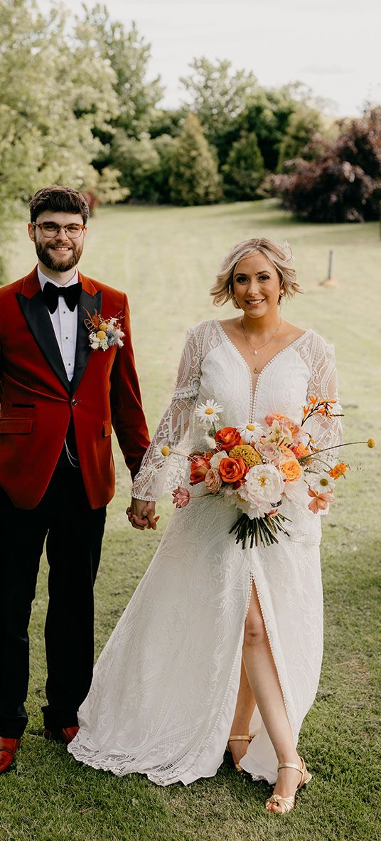 Bride holding an orange and yellow wedding flower bouquet in a boho dress with groom in an orange tux