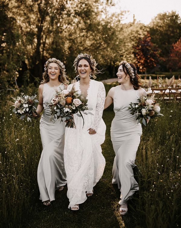 Bridesmaids in silver satin dresses and bride in a lace dress walking through the fields with a dried flower crown on their heads