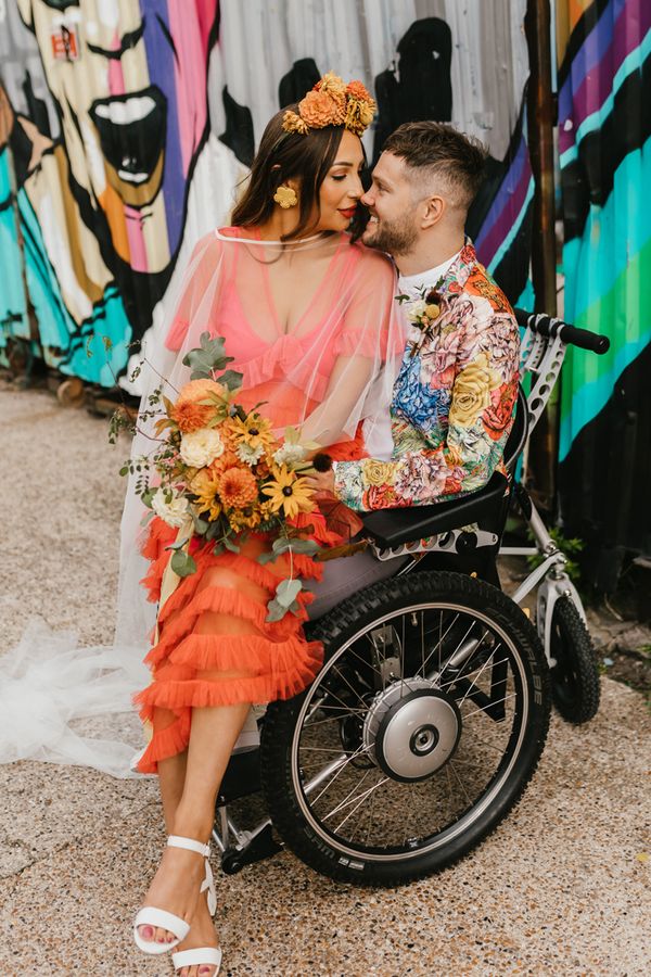 Bride in a coral wedding dress sitting on her grooms lap in a wheelchair for brightly coloured intimate elopement