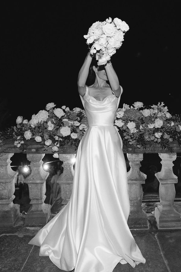 Black and white photo of a bride in a floor-length wedding dress holding a bouquet raised in the air.