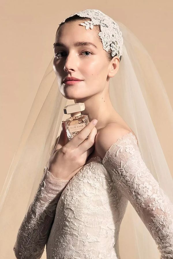 Bride wearing a lace wedding dress and veil, holding a bottle of Elie Saab Le Parfum Bridal Eau de Parfum Bridal Fragrance against a soft beige background - Wedding Perfume