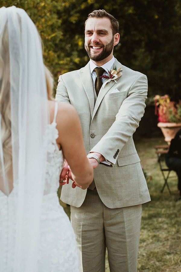 Groom wears classic stone beige linen suit, white shirt and wool green tie alongside a floral buttonhole