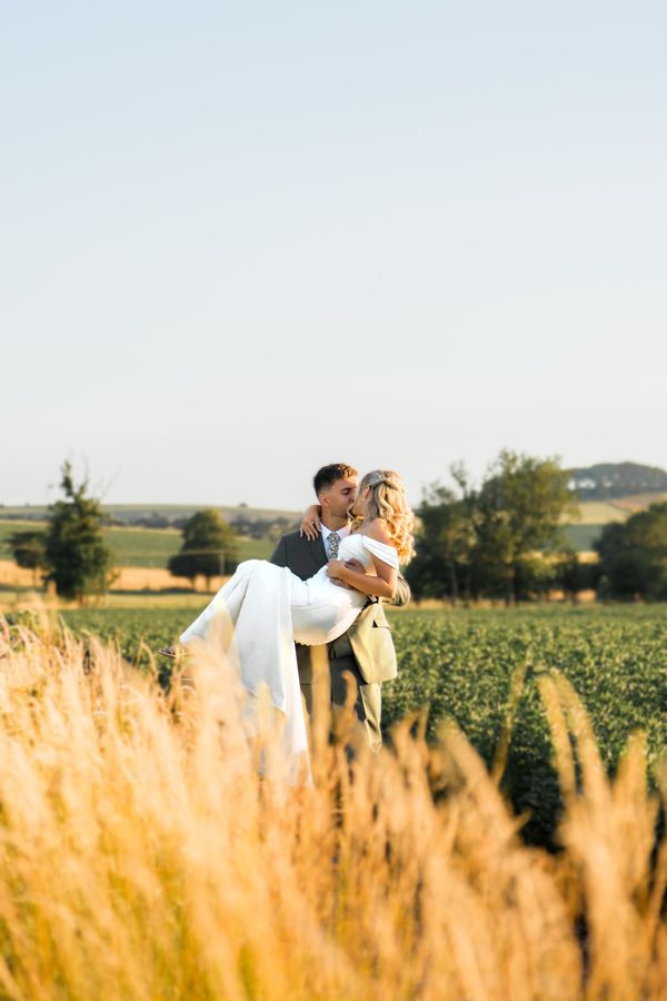 A romantic golden hour portrait of a groom sweeping his bride into his arms in a sunlit countryside meadow, framed by tall golden grass by Alex Gell Photography.