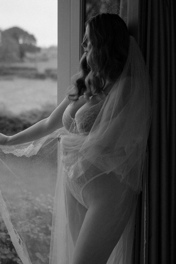 Bridal boudoir portrait of a bride wearing delicate ivory lace lingerie and a cathedral-length veil, standing beside a floor-to-ceiling window overlooking the Hampshire countryside. Captured in soft natural light with a fine art, black and white aesthetic. Photography by Rebecca Frost Photography.