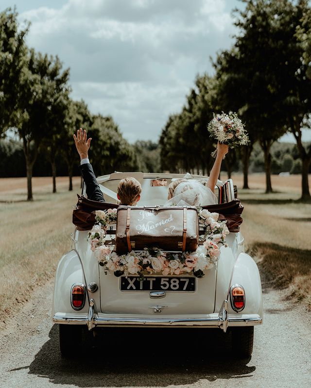 Bride and groom ride away in their wedding car with an old fashioned suitcase and flower decor.