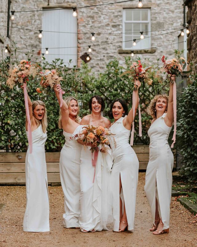 Bride in Alexandra Grecco dress with bridesmaids in white one shoulder dresses and neutral wedding bouquets.