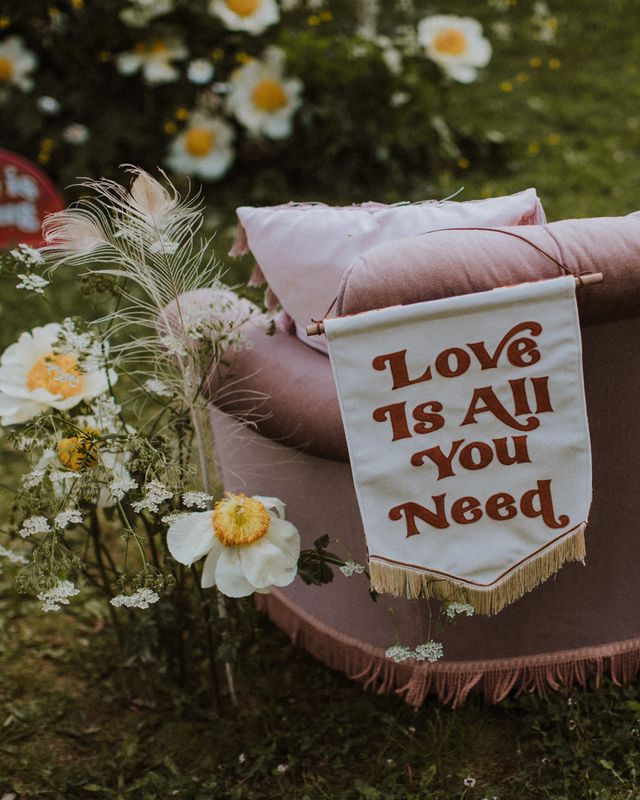 Love Is All You Need fabric banner resting on a velvet sofa surrounded by wildflowers for sixties wedding inspiration at a farm