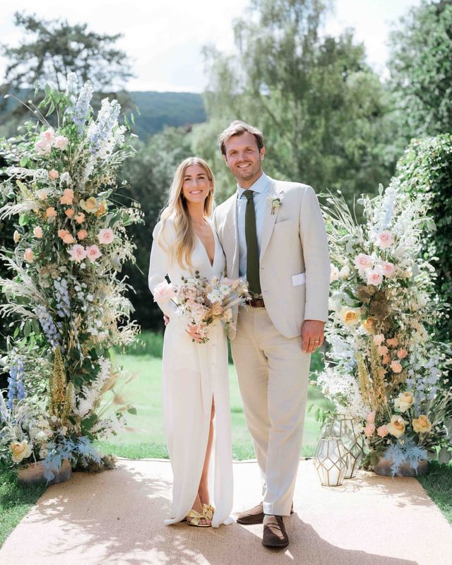 Bride wearing custom Bon Bride gown with plunging neckline and front slit with groom in cream suit in front of pastel flower displays for garden tipi wedding 