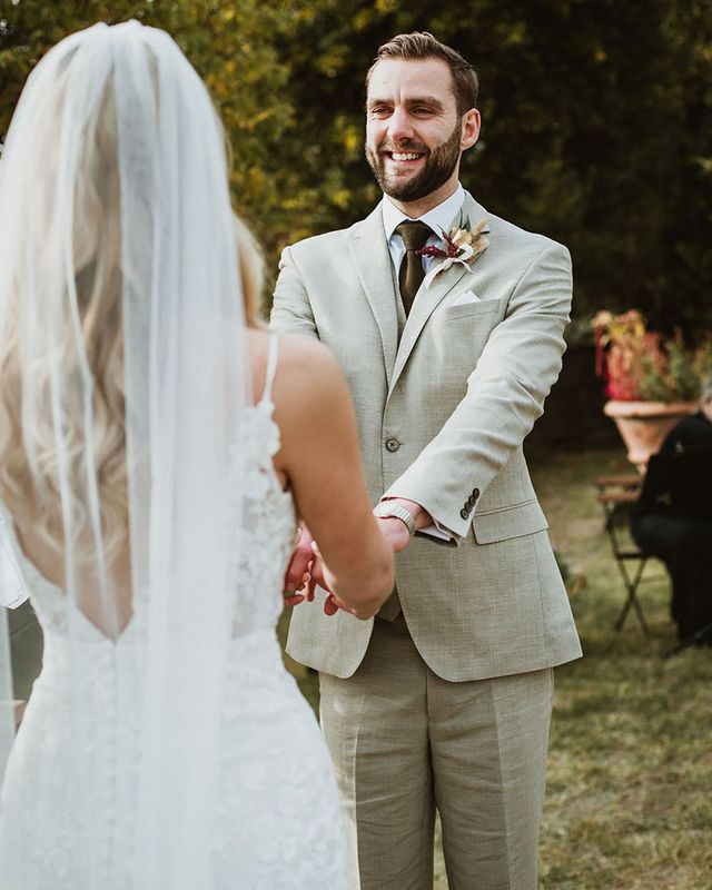 Groom wears classic stone beige linen suit, white shirt and wool green tie alongside a floral buttonhole