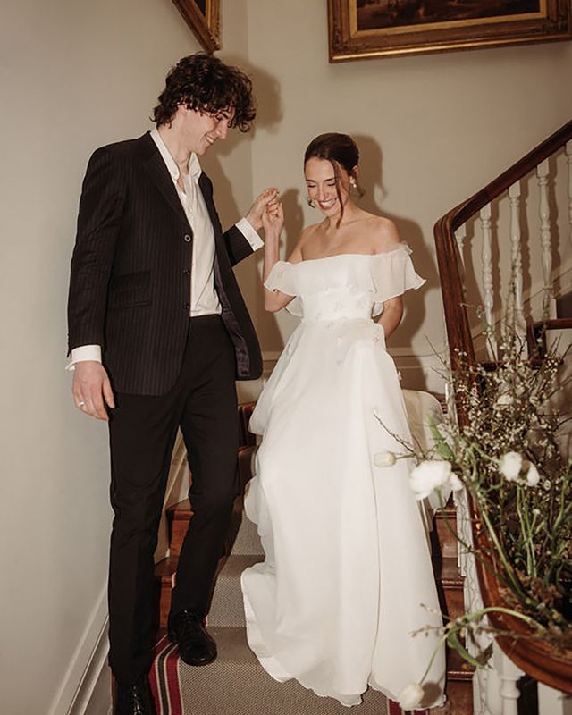 bride coming down the stairs with groom while wearing off-shoulder maxi wedding dress