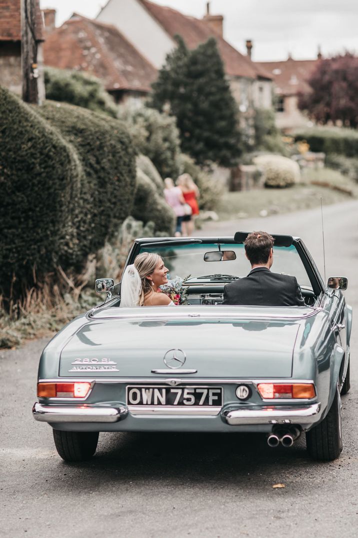 Bride wearing a wedding veil holding her pink and blue bouquet in the air driving off with her husband in a vintage blue convertible wedding car 