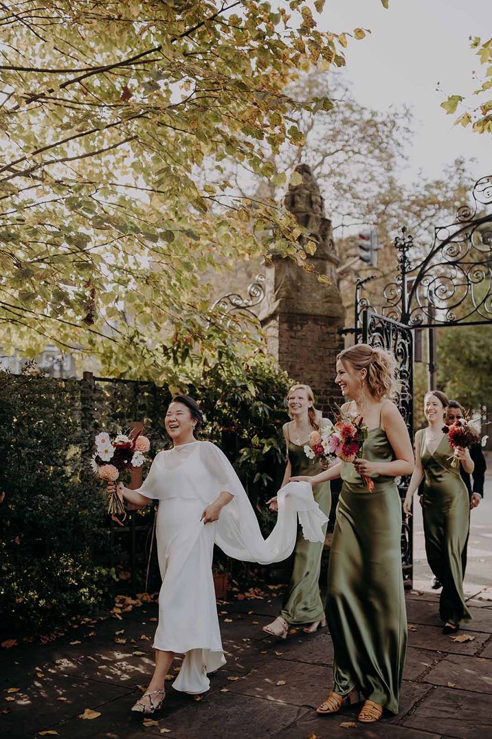 Bridesmaids in satin sage green bridesmaid dresses walk behind the bride and lift her wedding dress with a cape