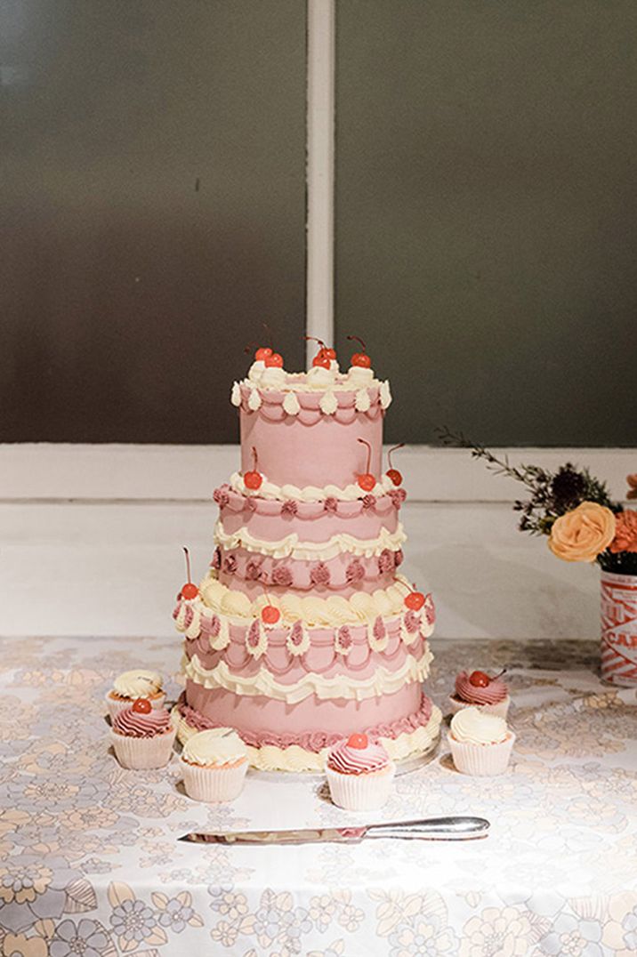 Three tier purple retro wedding cake with white icing topped with cherries surrounded by matching cupcakes for 60s style wedding 