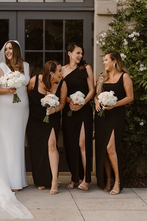 Bridesmaids wear long black bridesmaid dresses and hold white rose floral bouquets 