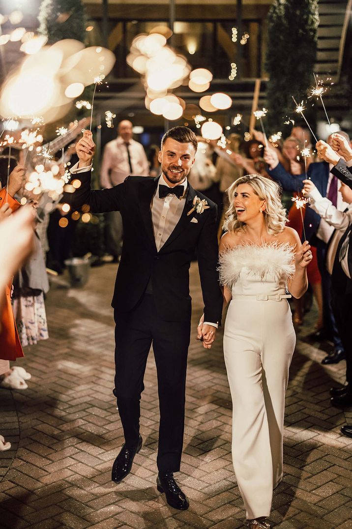 The bride in a Nadine Merabi wedding jumpsuit reception outfit has a sparkler send off with the groom in his black tuxedo 