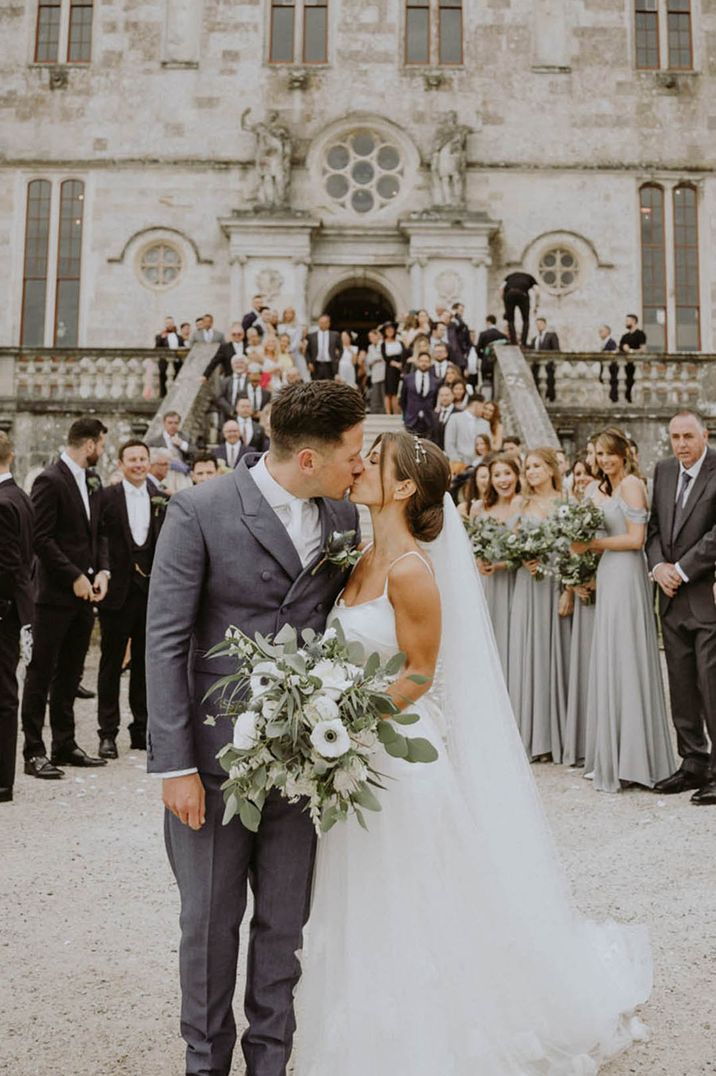 Groom in double breasted grey suit jacket kissing the bride in traditional wedding dress with sparkly hair piece and white bouquet with anemones 